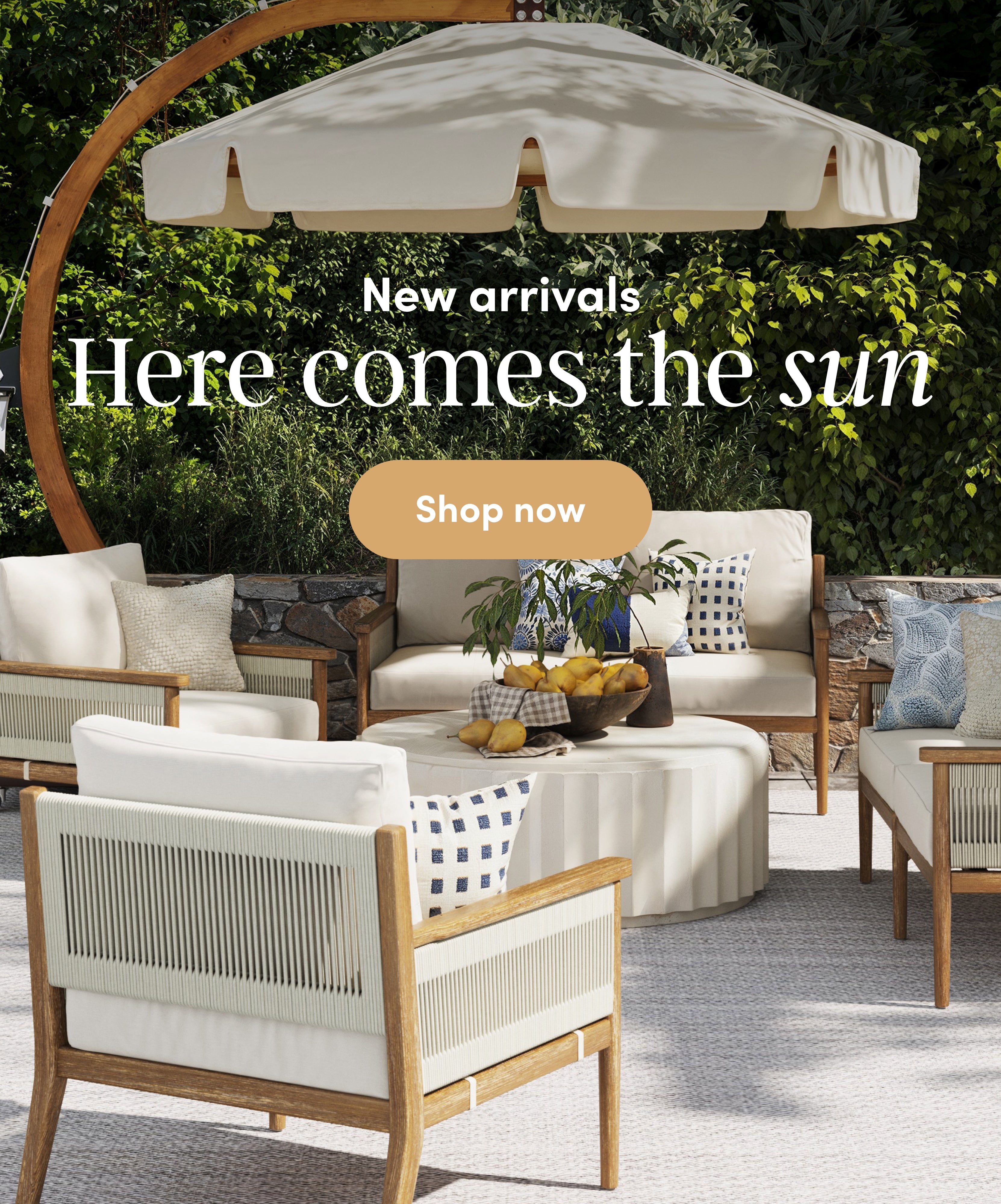 Outdoor patio furniture set with beige cushions and a round table under a large cream umbrella. Text reads: New arrivals. Here comes the sun. Shop now. Greenery and a stone wall are in the background.
