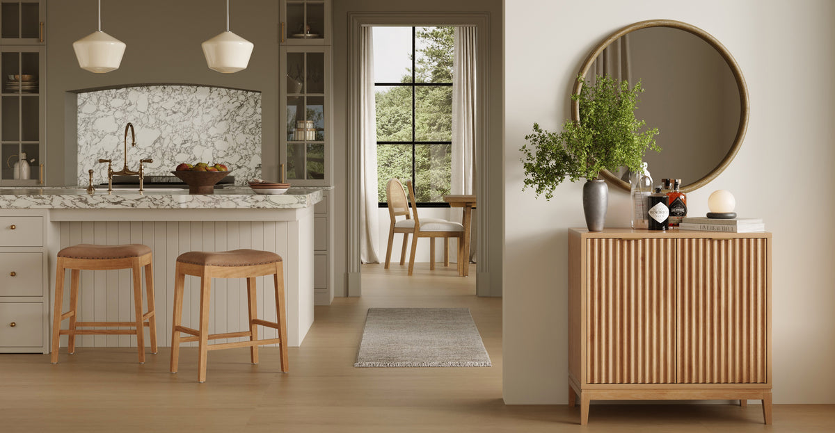 A modern kitchen with a marble-topped island and two stools, leading to a dining area with a view of trees, and a wooden console table with a round mirror in the foreground.