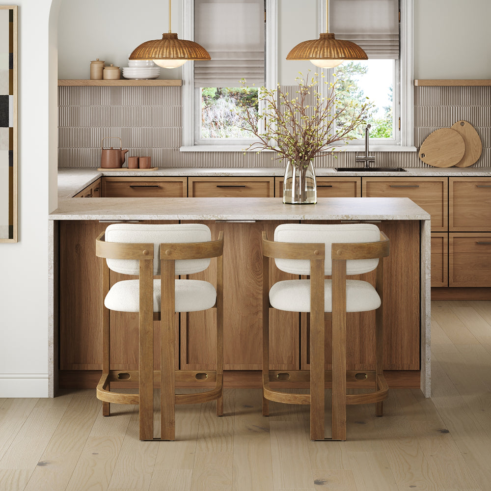 Two white upholstered wooden bar stools at a kitchen island with a light stone countertop, facing a wood-cabinet kitchen.