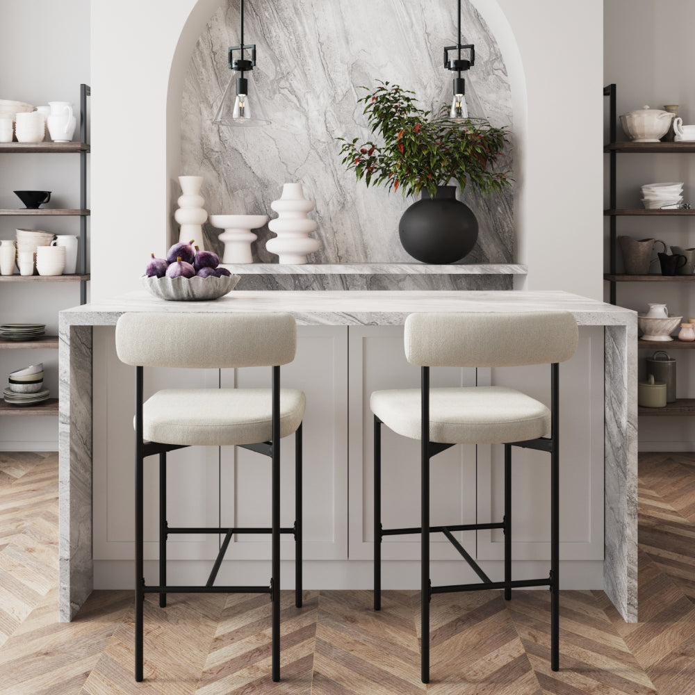A marble kitchen island with two cream-colored stools, a bowl of figs, and shelves filled with various ceramic dishes.