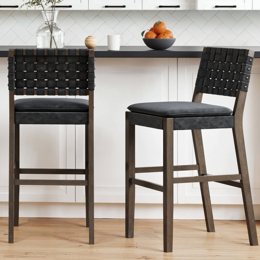 Two bar stools with dark wood frames, dark gray upholstered seats, and black woven leather-look backrests sit in a kitchen.