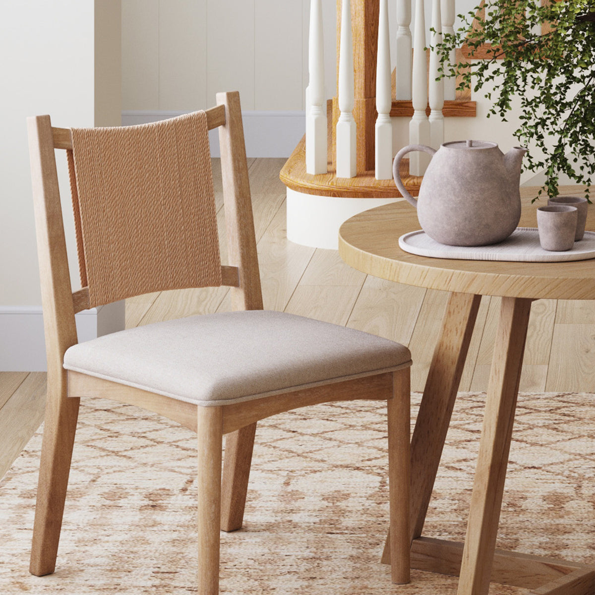 A wooden dining chair with a woven rope backrest and upholstered seat, positioned beside a round wooden table holding a teapot and two cups.