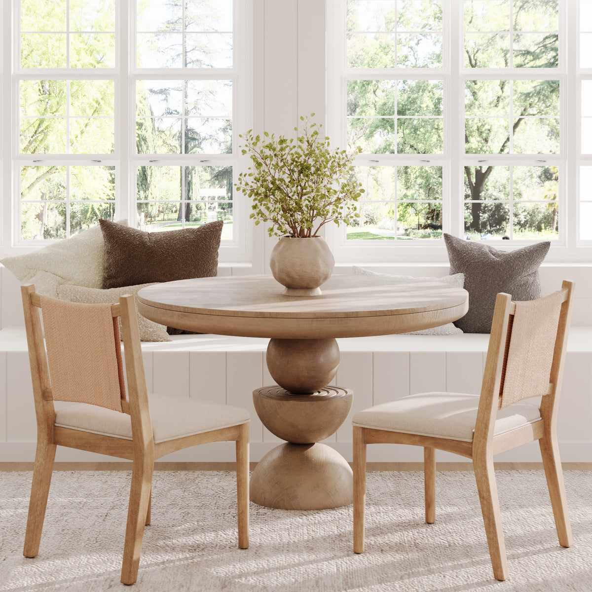 A round wooden pedestal table with two chairs, set in front of a windowed bench with throw pillows.