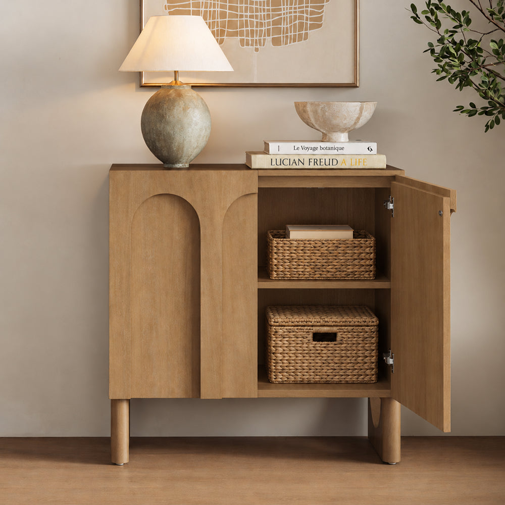 Wooden cabinet with an arched detail on the door and an open side displaying two woven baskets on shelves, topped with a lamp and stacked books.