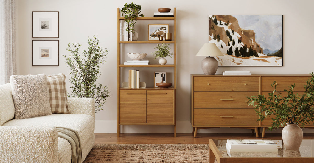 A living room scene featuring a light-colored sofa, a tall wooden bookshelf with decor, a wooden dresser topped with a lamp and artwork, and a glass-topped coffee table.