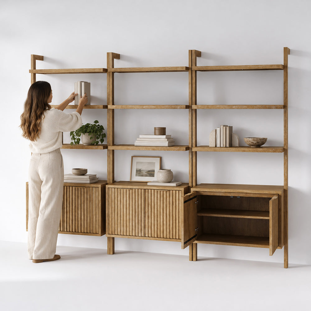 A person stands placing a book on a tall, modular wooden shelving unit that features open shelves and lower cabinets with slatted doors.