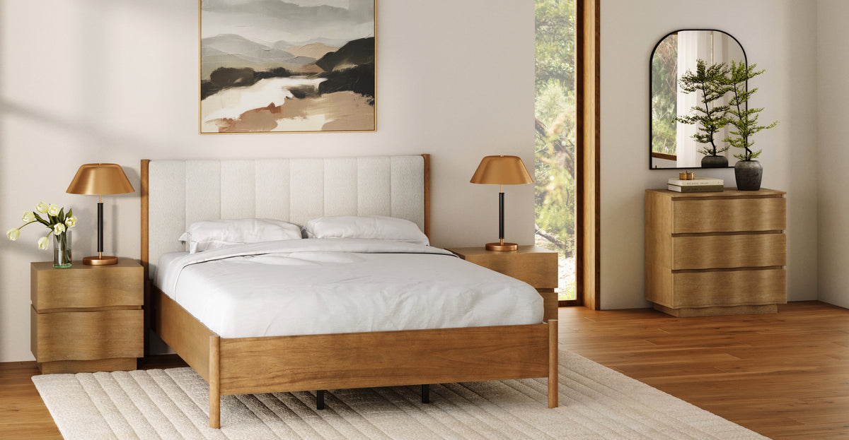 A bedroom with a wooden bed frame, upholstered headboard, two matching wooden nightstands with gold lamps, and a wooden dresser with a mirror and small potted plant.