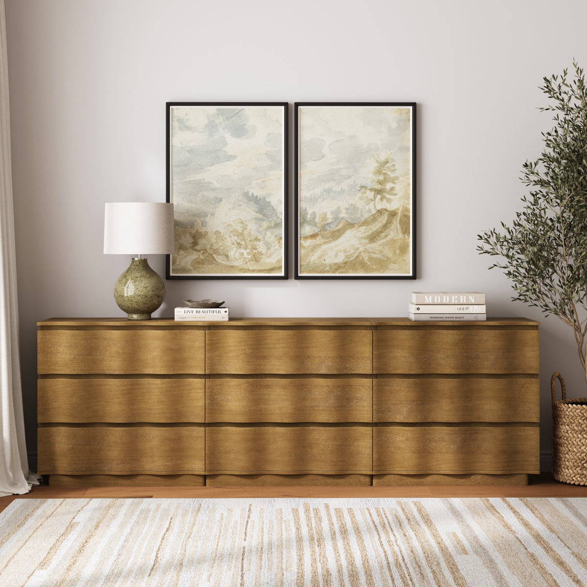 A wooden sideboard holds a lamp, two framed landscape prints, and stacks of books, with an olive tree in a basket to the right.