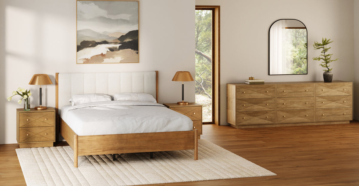 A bedroom with a wooden bed frame, two matching nightstands with lamps, and a long wooden dresser beneath an arched mirror, with a landscape painting on the wall.