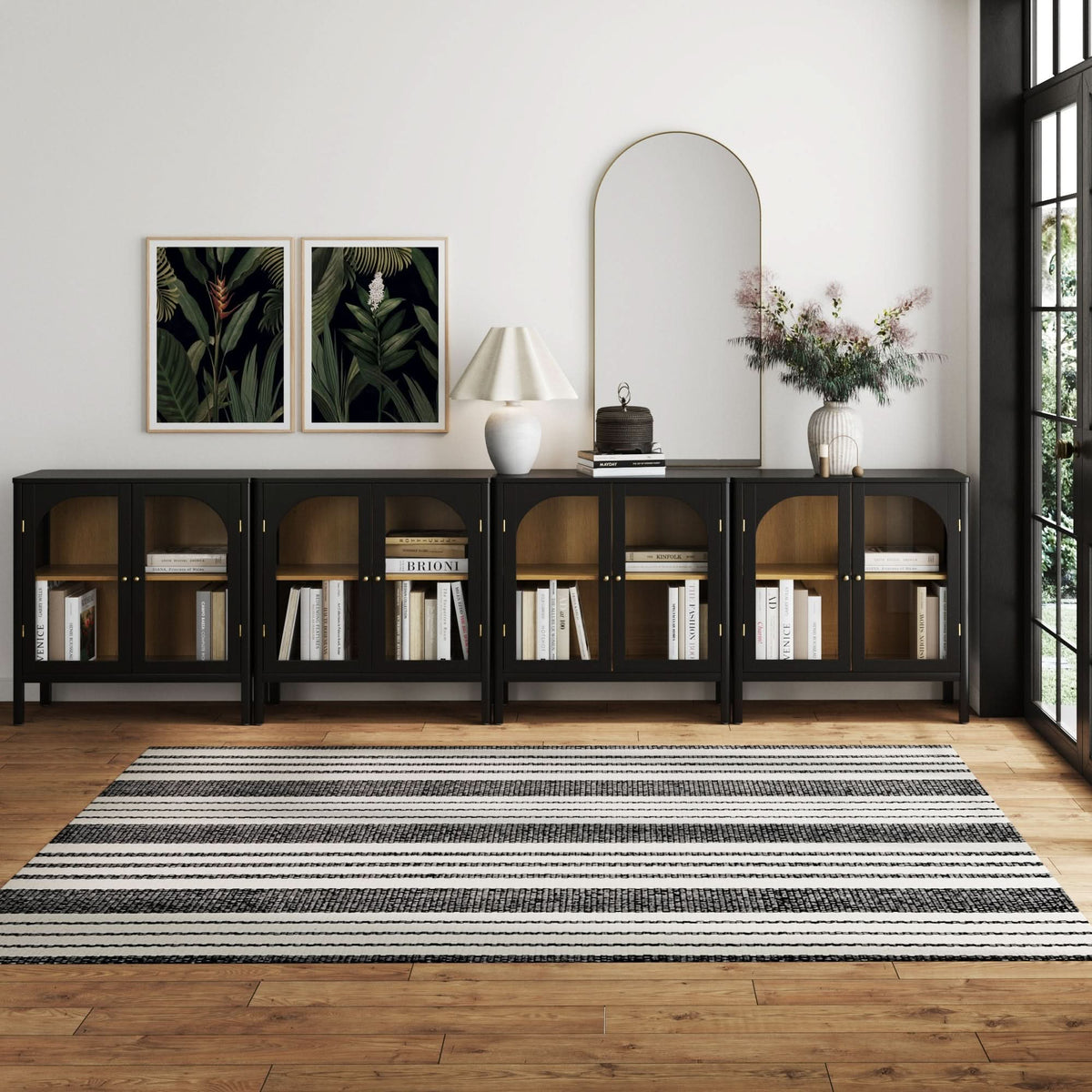 Four Nathan James Large Glass Door Curio Cabinet Credenza Black units arranged in a row, topped with a lamp, decorative items, and a mirror against a white wall with two framed botanical prints.
