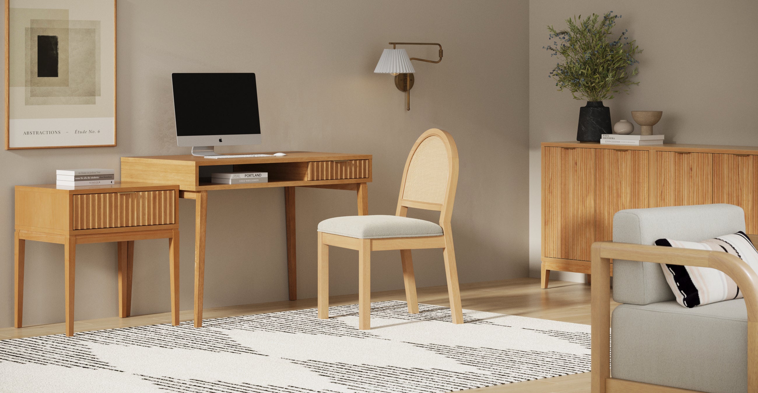 A modern home office with light wood furniture, including a desk, chair, cabinet, and side table. A computer sits on the desk; a rug, wall art, sconce, and vase with greenery add decor. Neutral colors create a calm atmosphere.