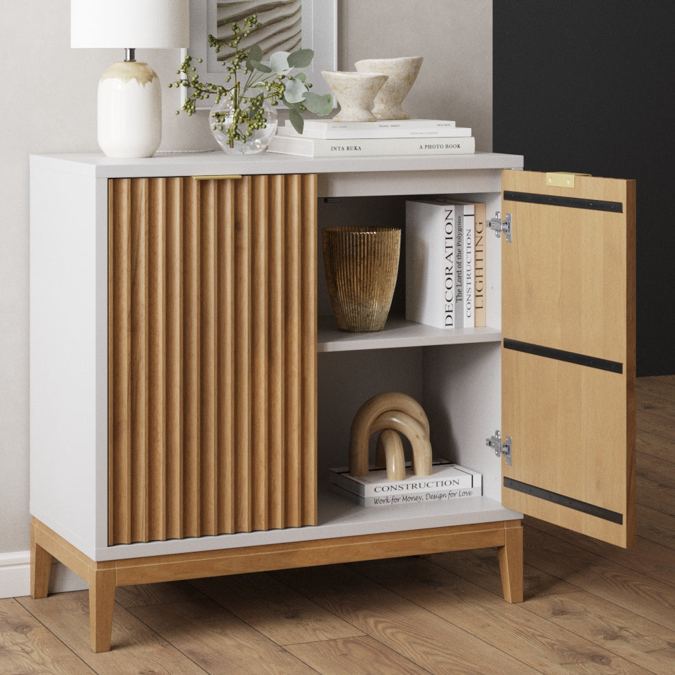 A modern wooden cabinet with one door open reveals shelves holding books and decorative objects. The cabinet has vertical slats on one side, a white top, and stands on four legs on a wooden floor.