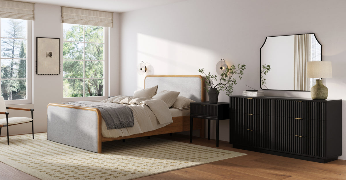 A bedroom featuring a light wood and grey fabric bed, a black nightstand, and a matching black fluted dresser topped with a mirror and lamp.