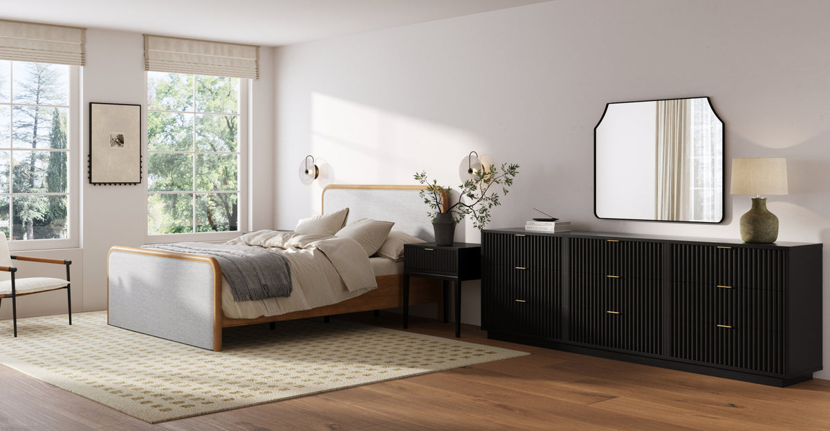 A bedroom featuring a bed with a light-colored headboard, a black nightstand with a plant, and a long black dresser with a matching mirror and lamp.