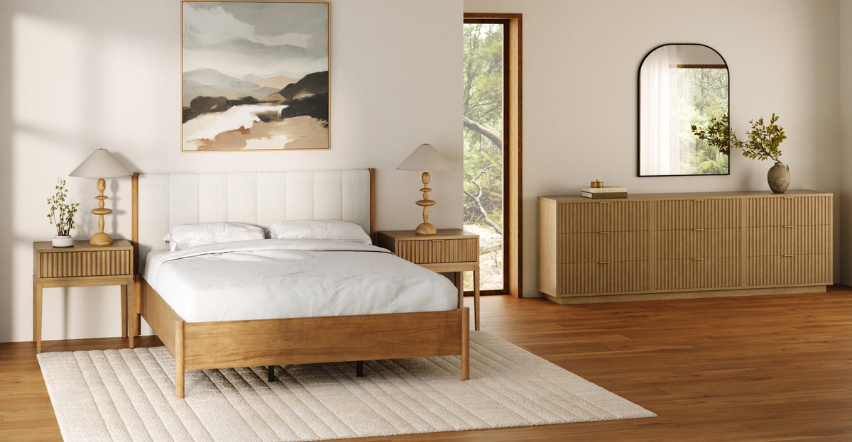 A bedroom featuring a wooden bed frame, two matching nightstands with lamps, and a fluted wooden dresser topped with a mirror and vase.