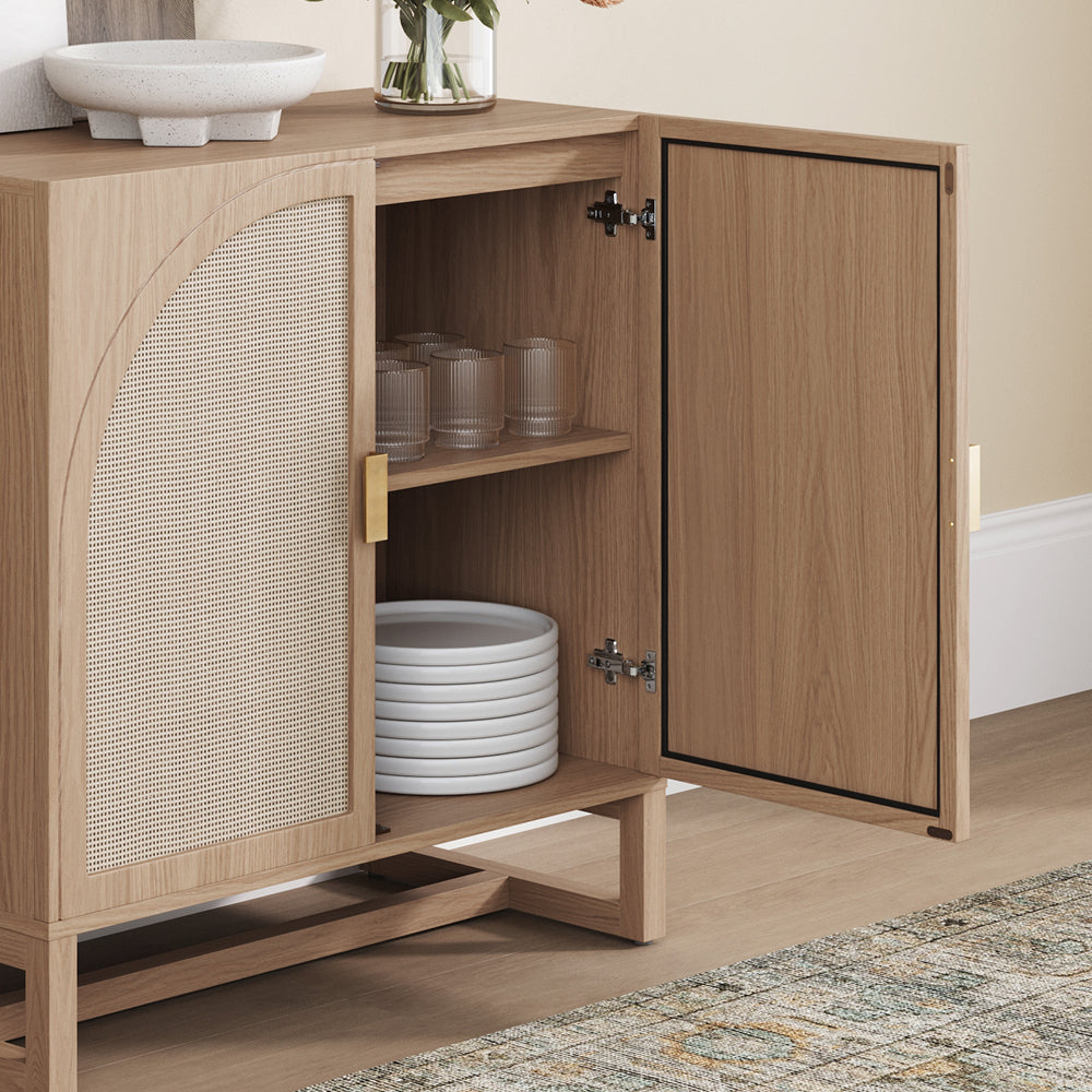 Light wood cabinet with a curved cane door panel, shown with the right cabinet door open to reveal a shelf holding glassware and stacked plates.