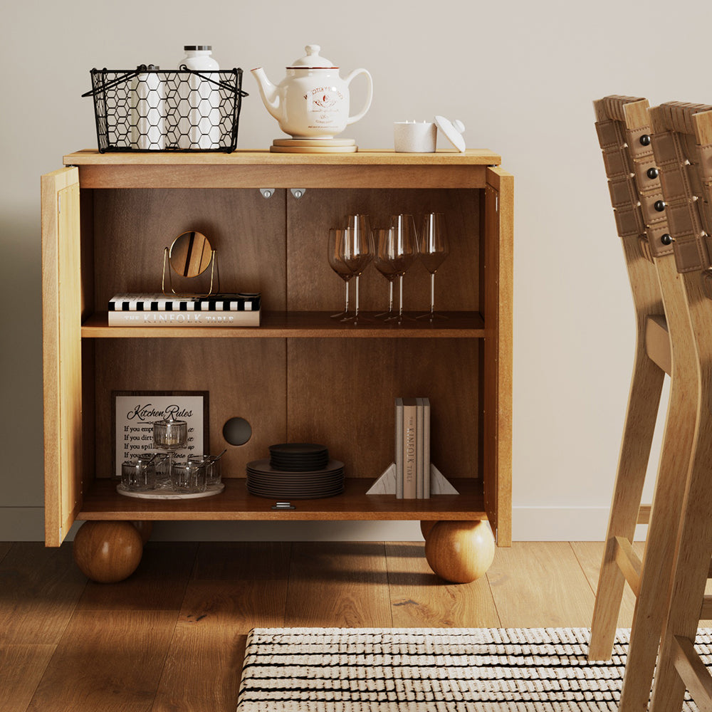 A wooden cabinet with open doors revealing shelves stocked with glassware, a stack of plates, and books, topped with a wire basket, teapot, and candle.