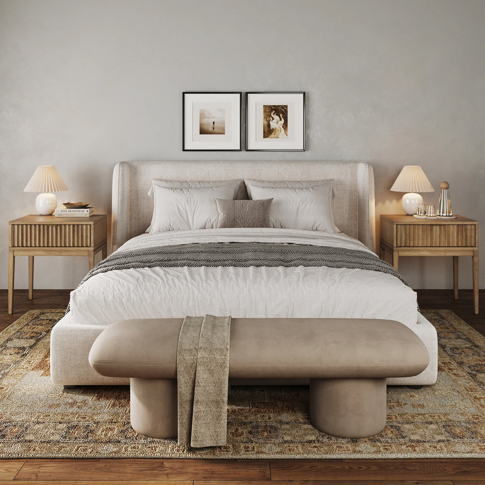 A bed with a cream-colored upholstered headboard, flanked by two wooden nightstands with lamps, sitting on a patterned rug with an oval bench at the foot of the bed.