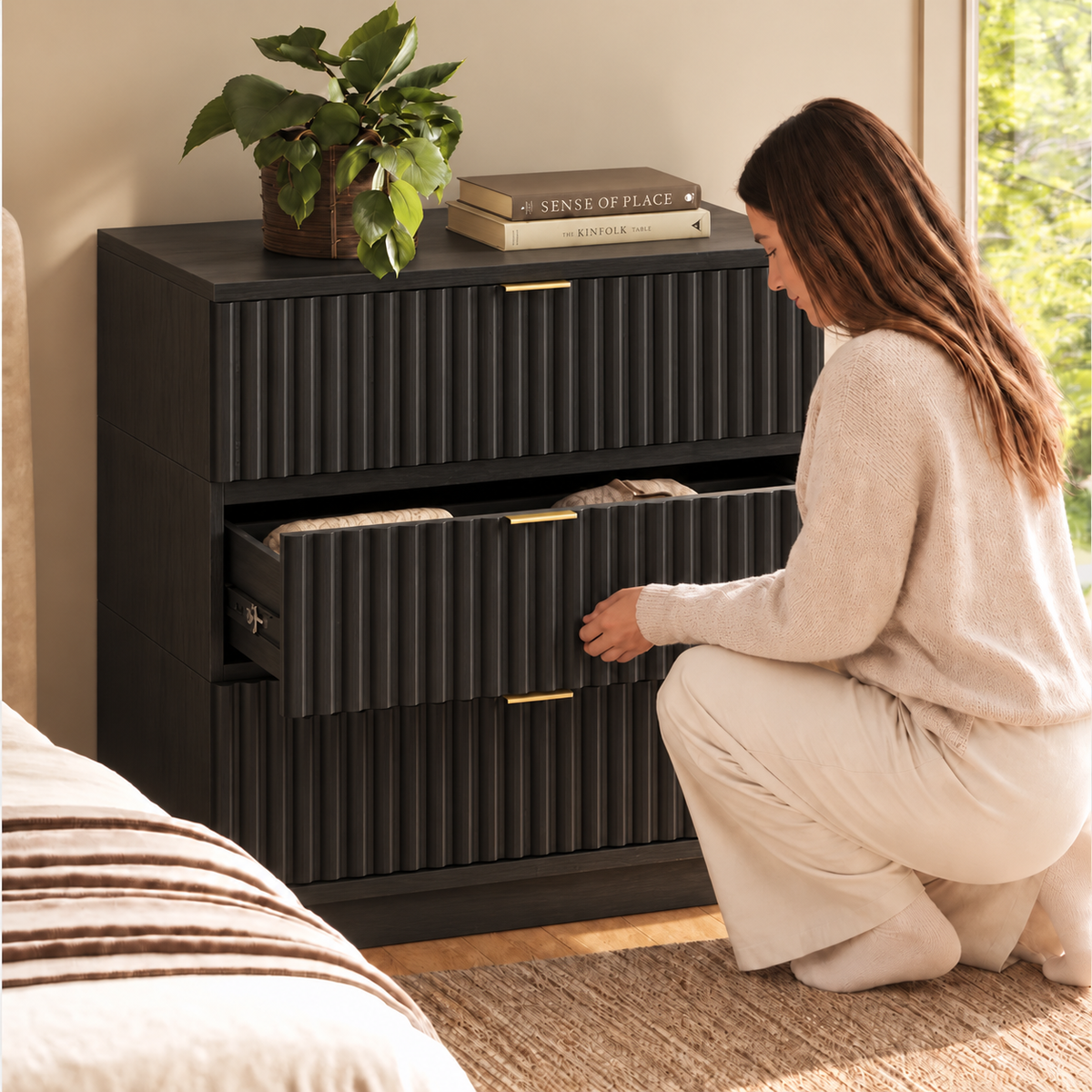 A woman in a cream sweater kneels beside a black three-drawer dresser, pulling open the middle drawer.