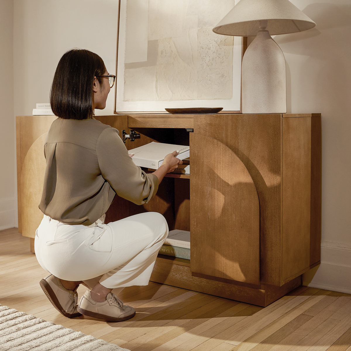A woman in a green blouse and white trousers crouches to place a book inside a wooden cabinet.