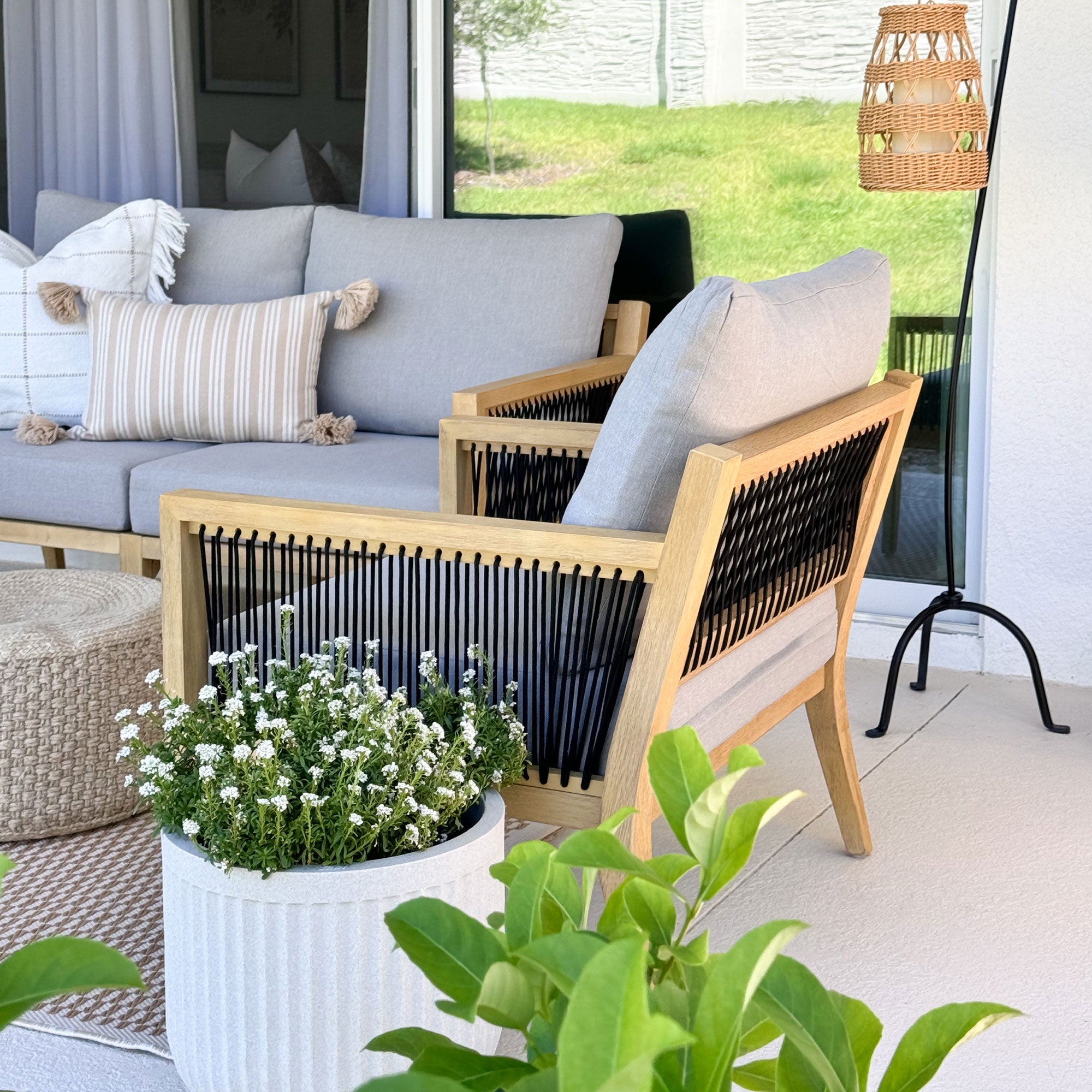 A modern patio with a light wood and black rope accent chair, gray cushions, and a matching sofa with striped and textured pillows. A large white planter with white flowers sits in front, and greenery is outside the sliding door.