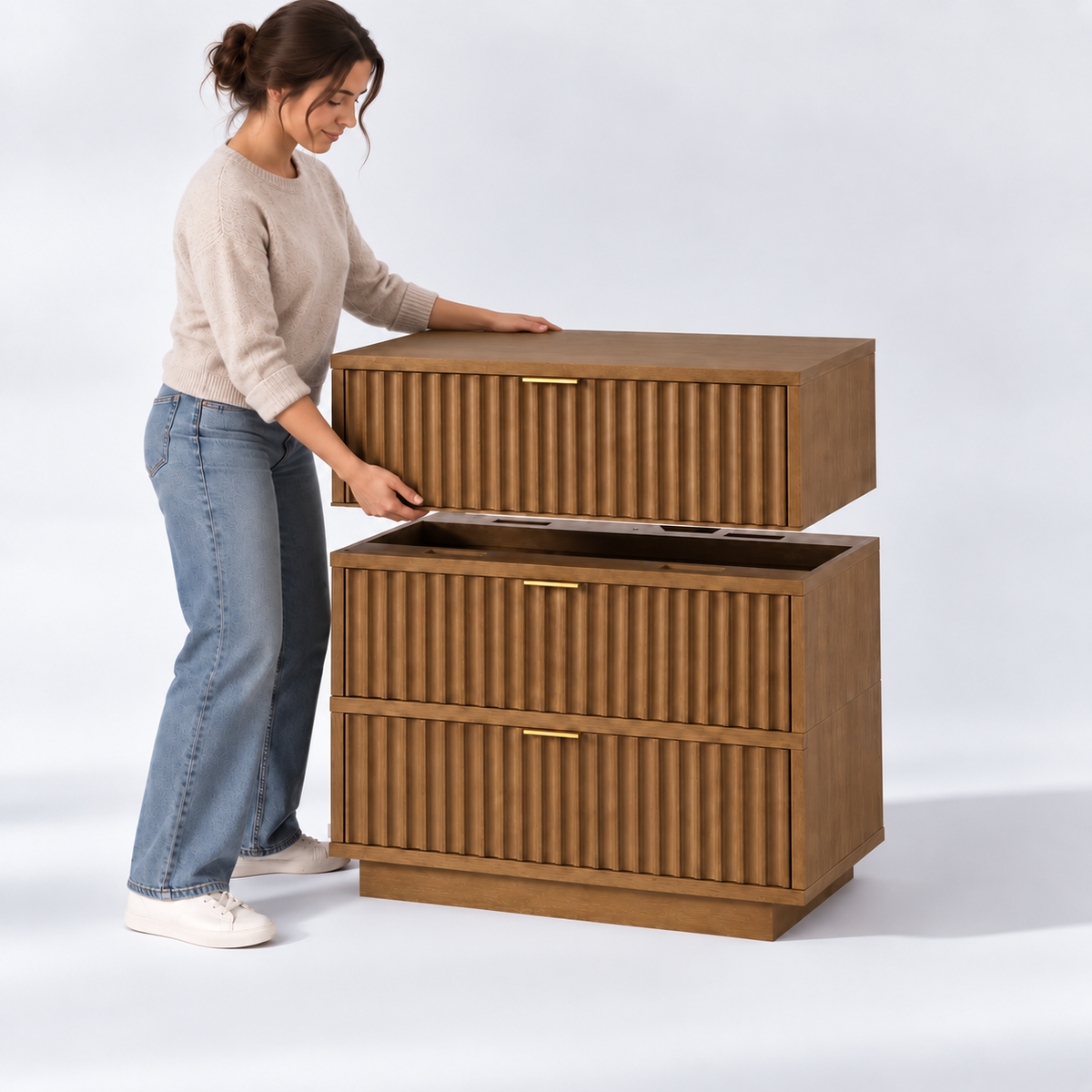 A person lifts the top drawer section off a three-drawer wooden dresser with a fluted front design and gold hardware.
