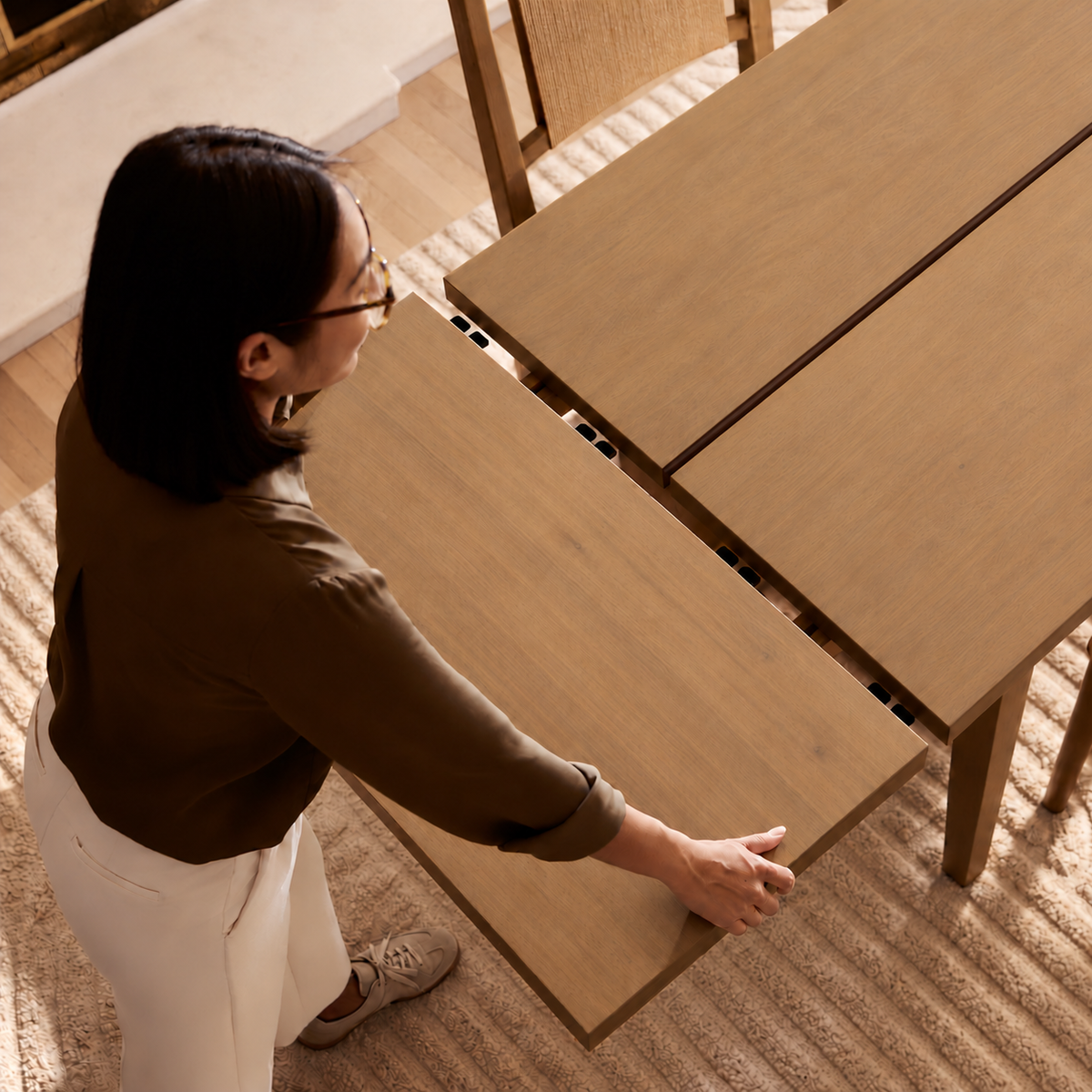 A woman standing at a light wood dining table, sliding an extension leaf into place.
