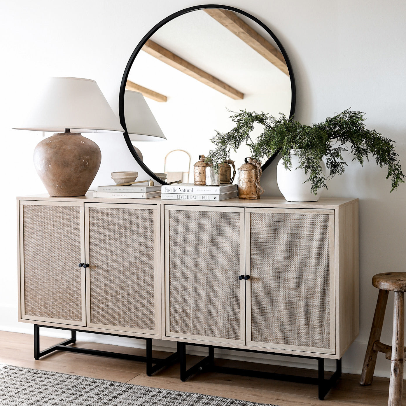 A light wood sideboard with woven doors sits against a white wall, topped with a large round mirror, ceramic lamp, green plant, stacked books, and decorative jars. A wooden stool sits beside the sideboard.