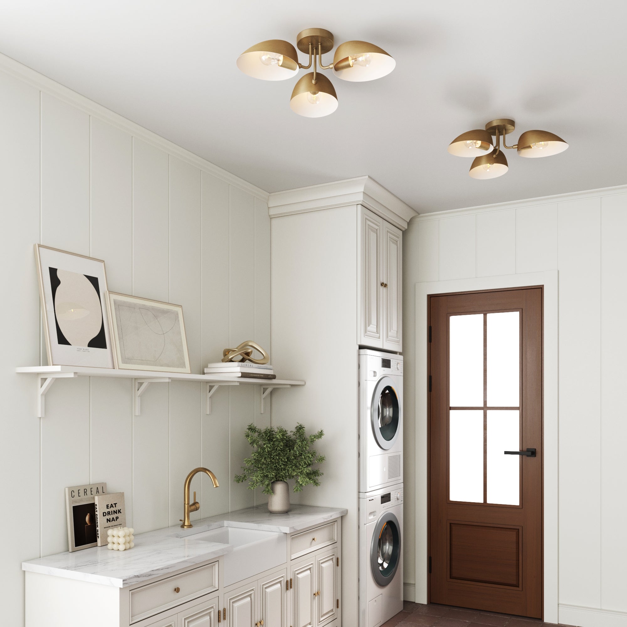 A bright laundry room with white cabinets, a stacked washer and dryer, a brown door with a glass window, gold light fixtures on the ceiling, and minimalist art on a shelf above a white sink with a gold faucet.
