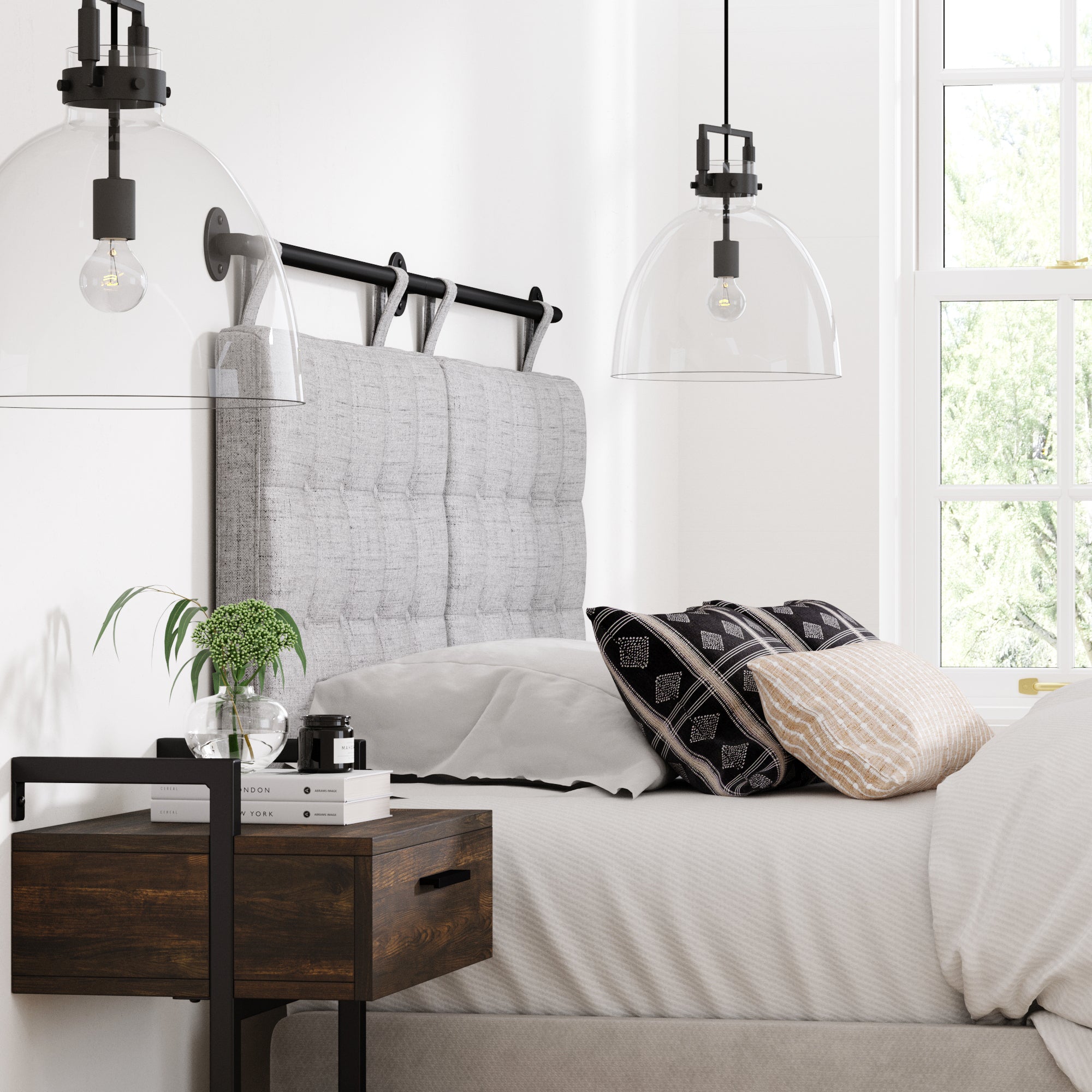 A cozy, modern bedroom with a gray upholstered headboard, patterned pillows, white bedding, a wooden nightstand with books and a plant, and two glass pendant lights. Sunlight enters through a window with a view of greenery.