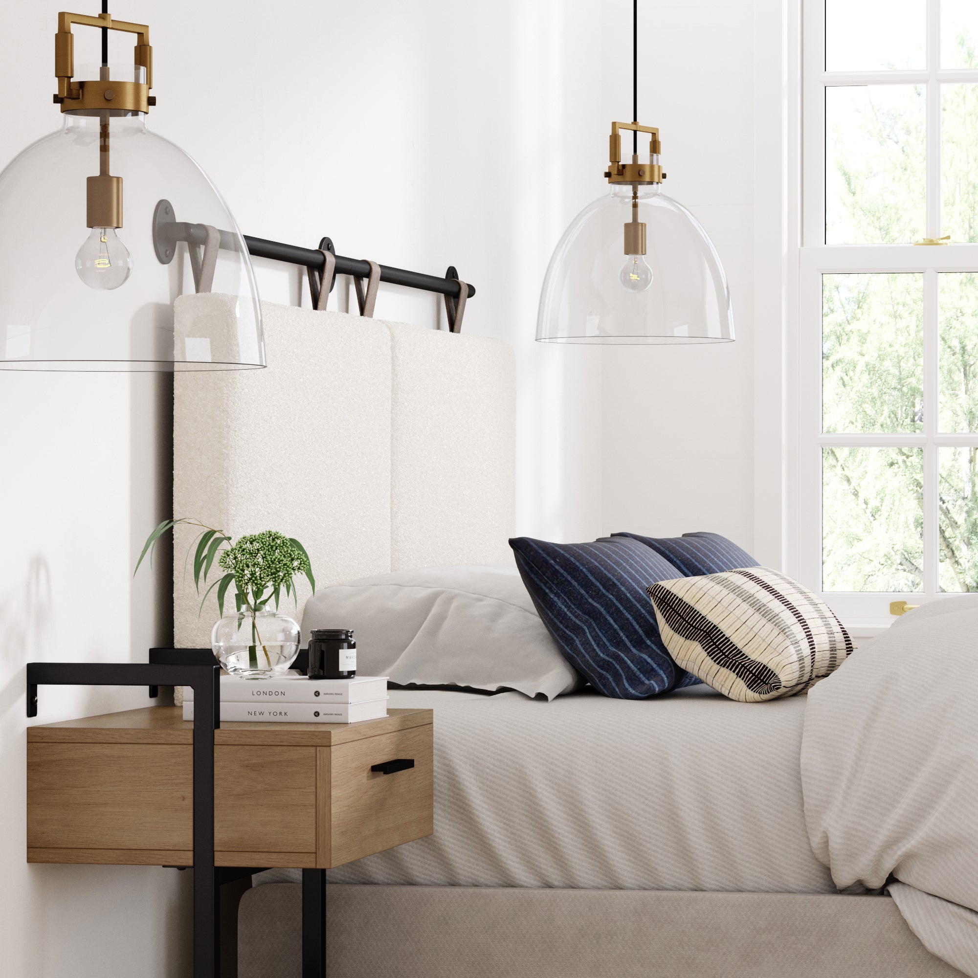 A modern bedroom with a beige bed, navy and plaid pillows, a wooden nightstand with books and a plant, and two glass pendant lights. Sunlight streams through a window, highlighting minimalist decor.