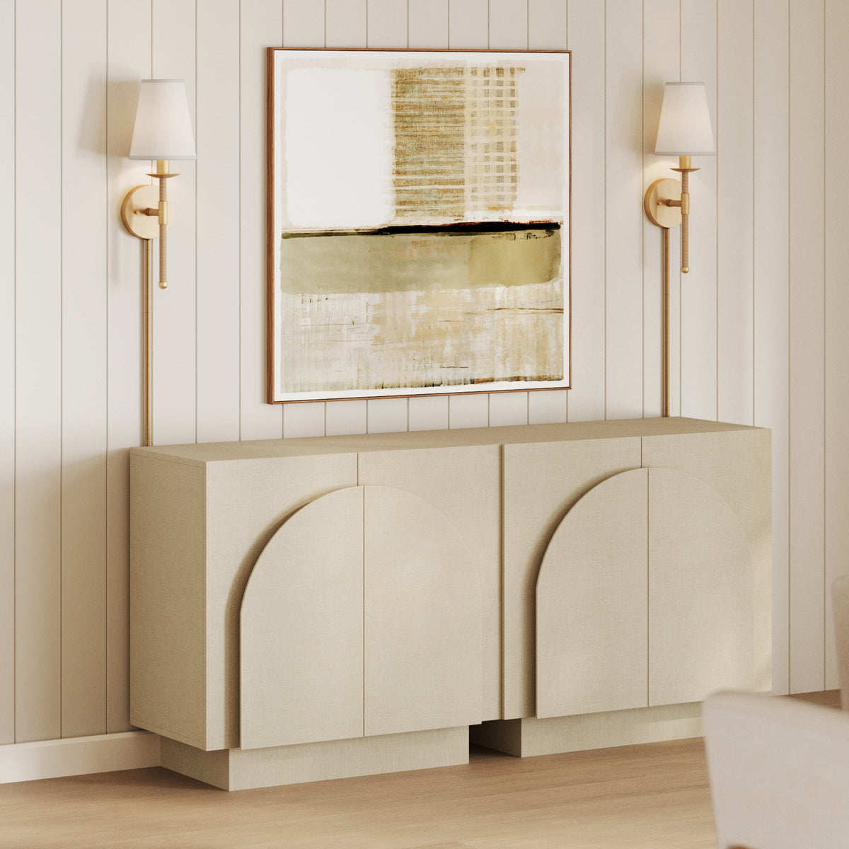 A light-colored sideboard with arched door details sits below a large abstract print and two gold wall sconces against a white paneled wall.