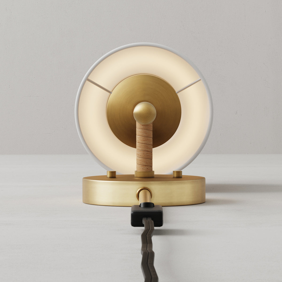 Round brass table lamp with a circular glowing light element and a wrapped cord detail on a flat base.