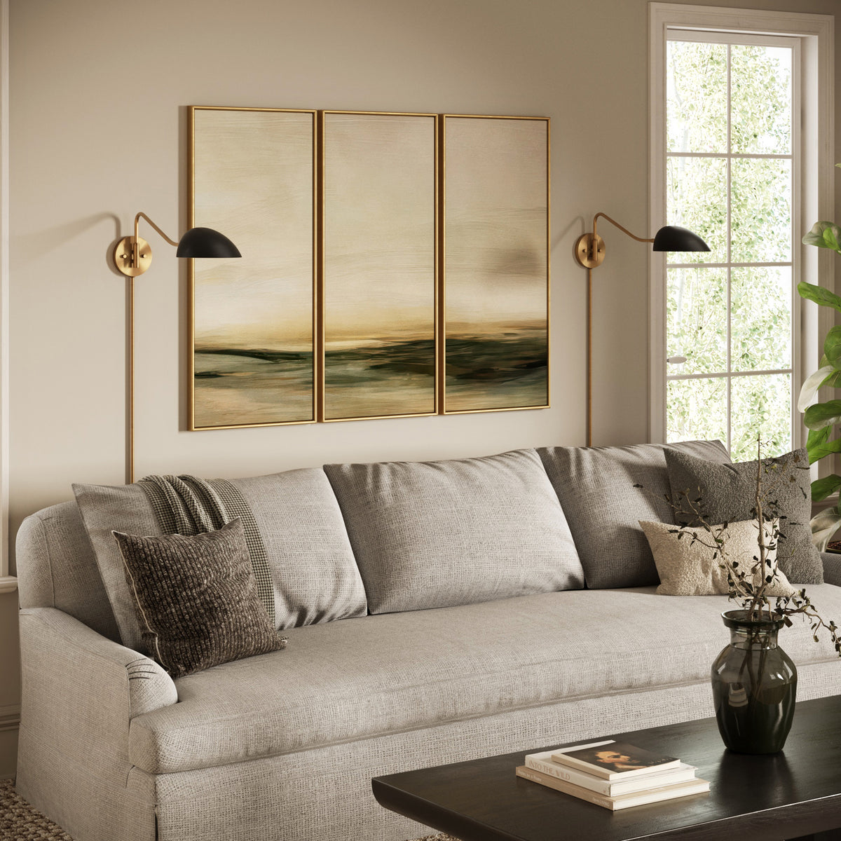A light gray sofa sits beneath three landscape paintings in gold frames, flanked by two brass wall sconces.
