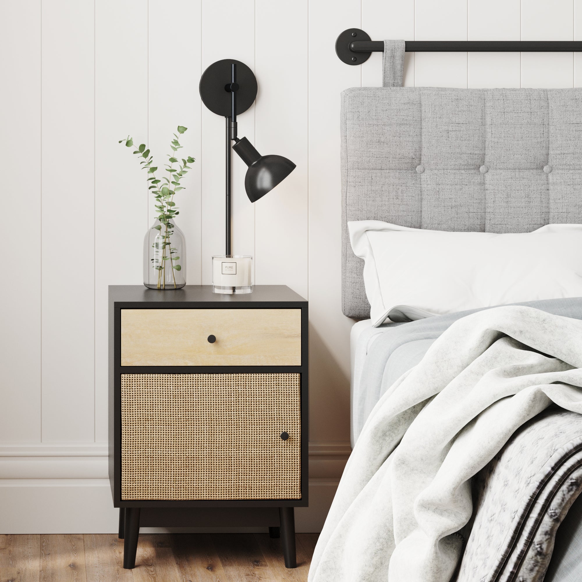 A modern bedroom scene with a gray upholstered bed, white bedding, and a black nightstand holding a glass vase with greenery, a small candle, and a black wall-mounted reading lamp above.