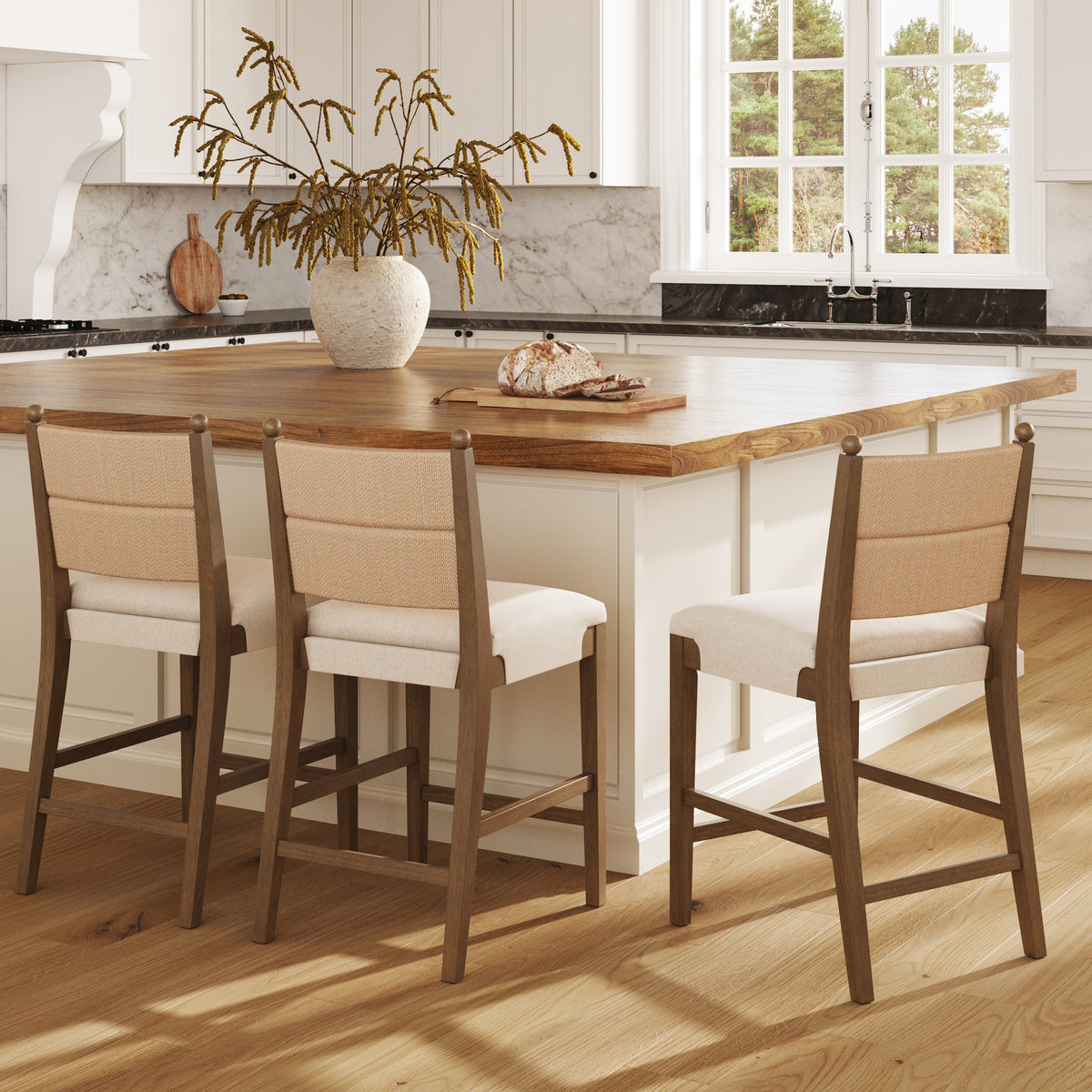 Three wooden bar stools with tan fabric seats and backrests sit in front of a kitchen island topped with a loaf of bread and a decorative vase of dried branches.