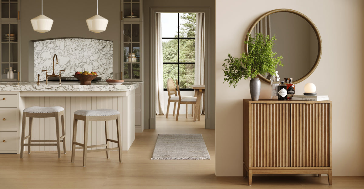 A kitchen with a marble-topped island and two stools, viewed through an open doorway near a wooden sideboard topped with a round mirror, a plant, and bottles.
