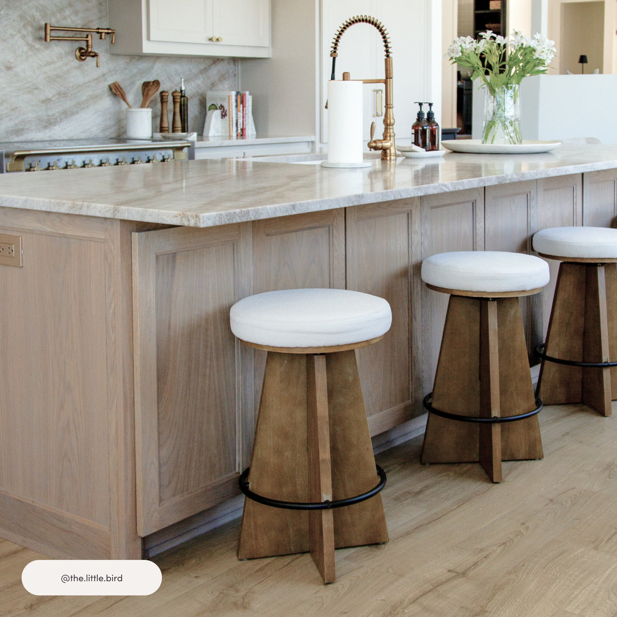 Three Nathan James Modern Backless Swivel Boucle Counter Stools with white upholstered seats and wooden pedestal bases with black footrests, arranged along a kitchen island.