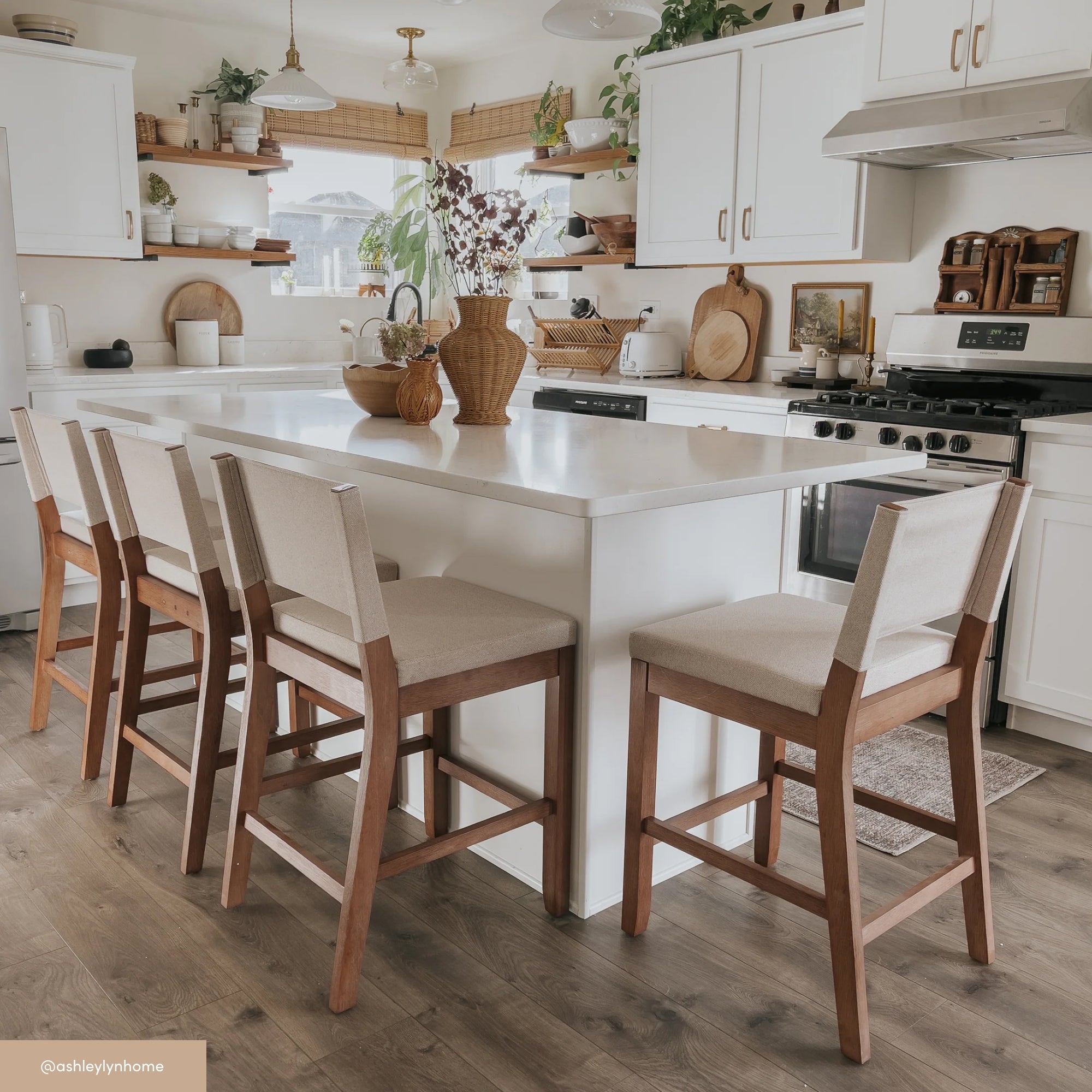 A bright, modern kitchen with white cabinets, a large white island, four cushioned wooden chairs, plants, wooden accents, and natural light streaming through the windows.