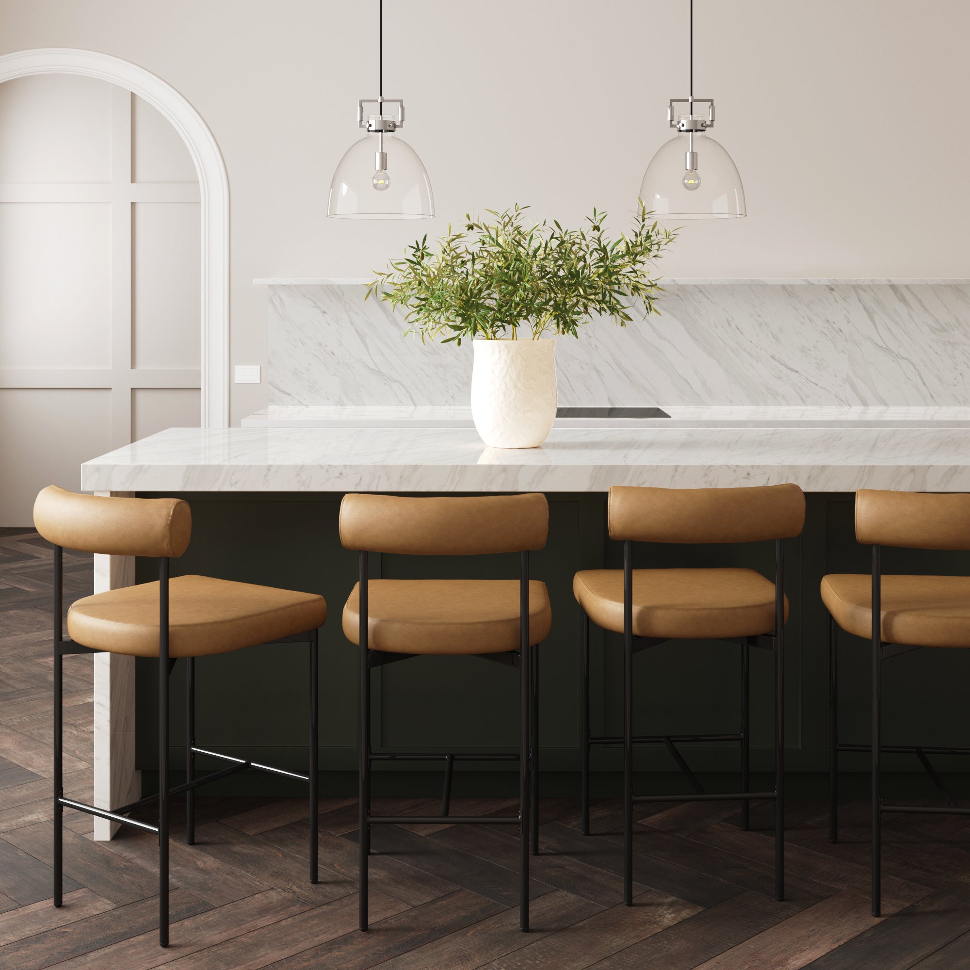 A modern kitchen with a marble island, four tan leather bar stools with black legs, a white vase with green foliage, pendant lights above, and dark herringbone wood flooring.