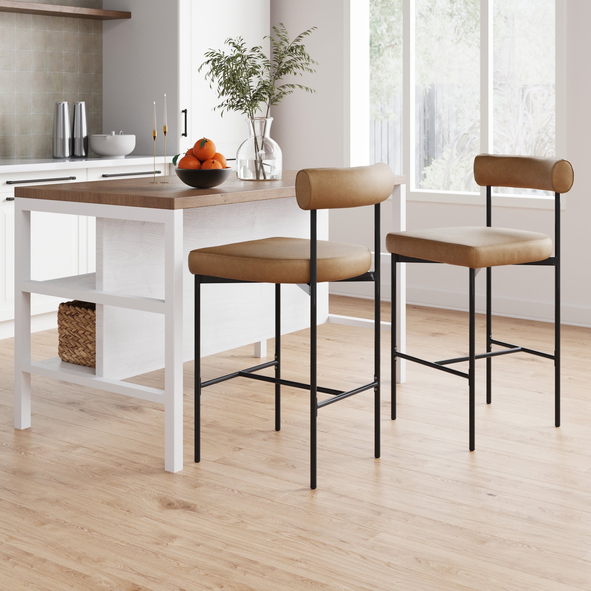 A modern kitchen with a white island, two tan and black barstools, a bowl of oranges, a plant in a vase, and large windows letting in natural light onto a wood floor.