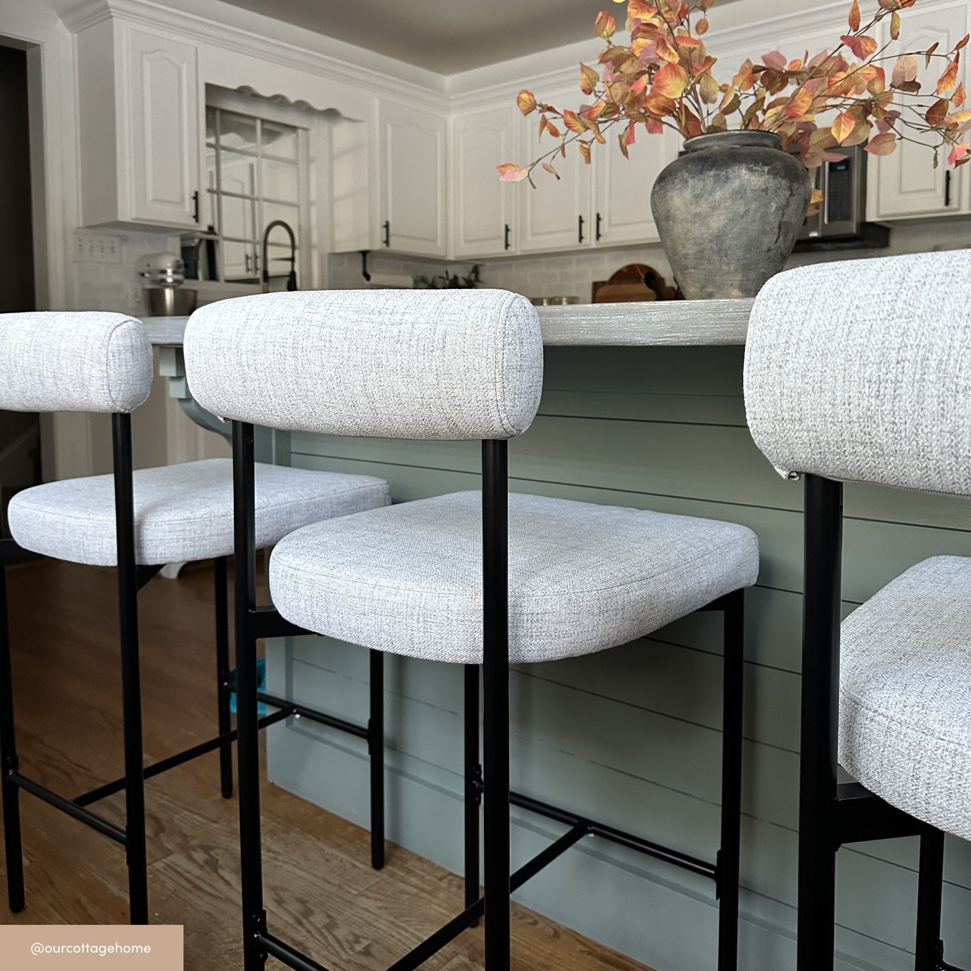 Three modern barstools with light gray upholstered seats and black metal legs are lined up at a kitchen island with a green base. A vase with autumn leaves sits on the counter; white cabinets are visible in the background.