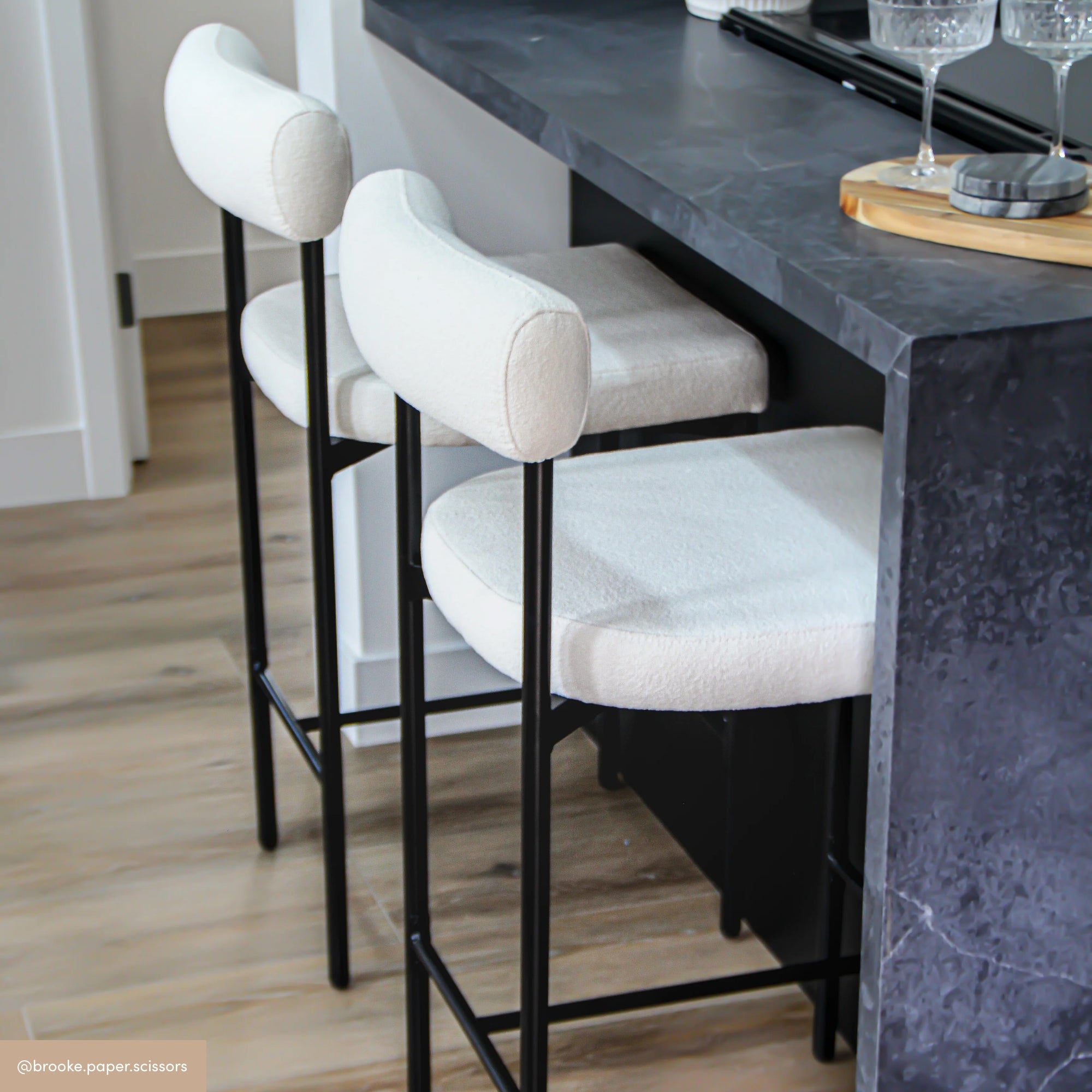 Two modern white upholstered bar stools with black metal legs are placed at a dark stone kitchen island. The countertop holds glassware and a wooden tray. The floor is light wood.