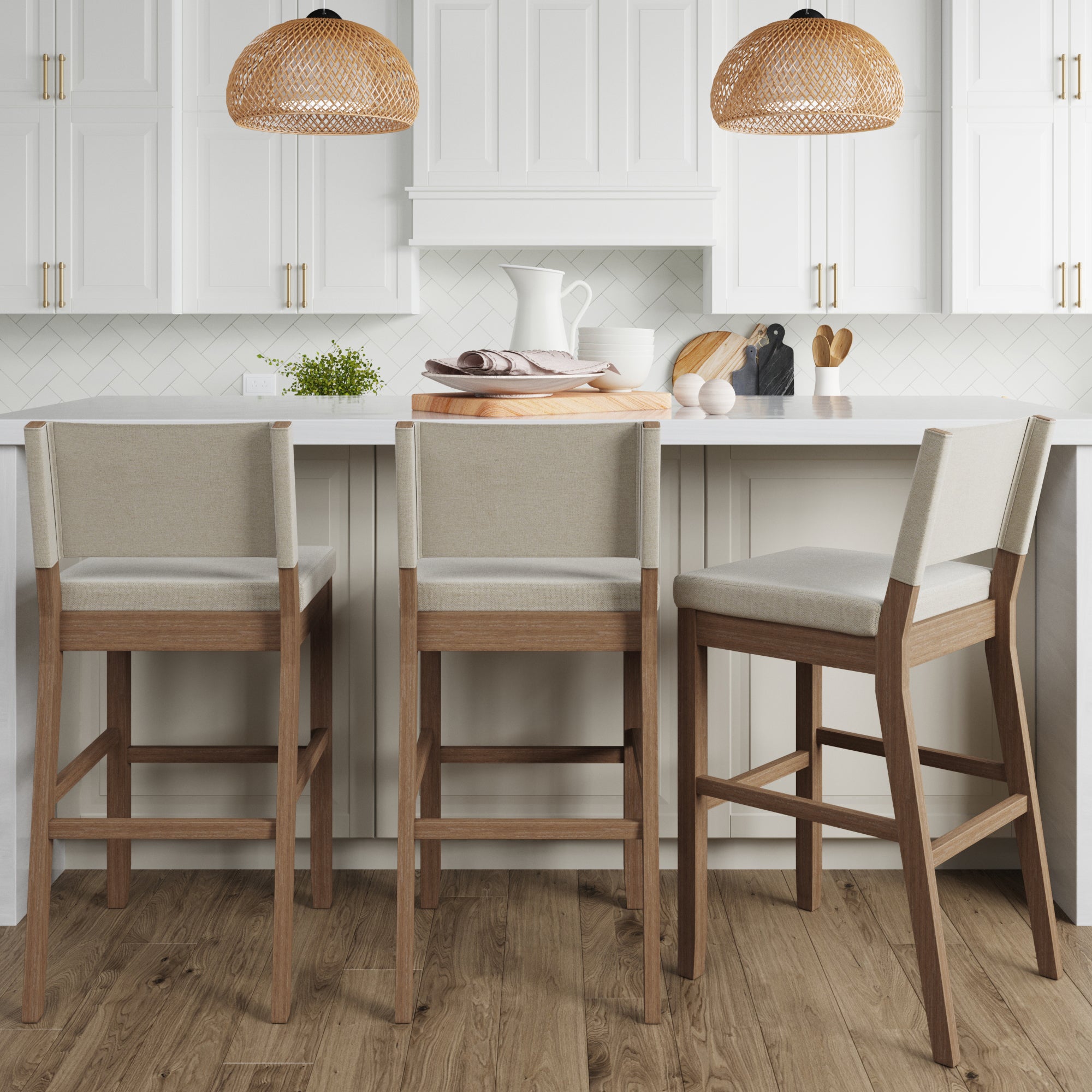 Three modern wooden bar stools with light beige cushions sit at a white kitchen island. The kitchen features white cabinets, a herringbone backsplash, wood floor, and two woven pendant lights. Kitchenware decorates the counter.