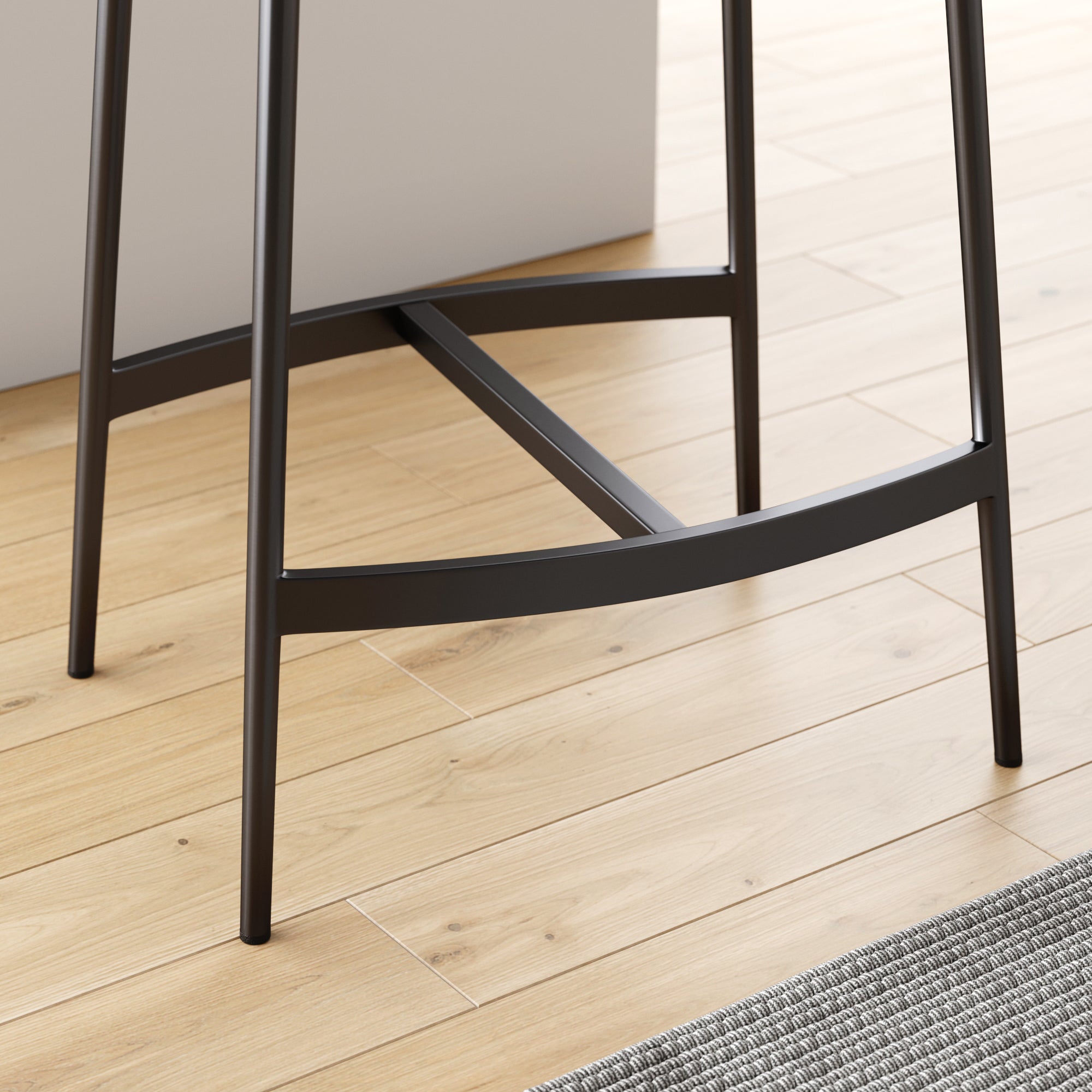 Close-up of black metal legs and footrest of a modern bar stool on light wooden flooring, next to a gray woven rug and a white countertop.