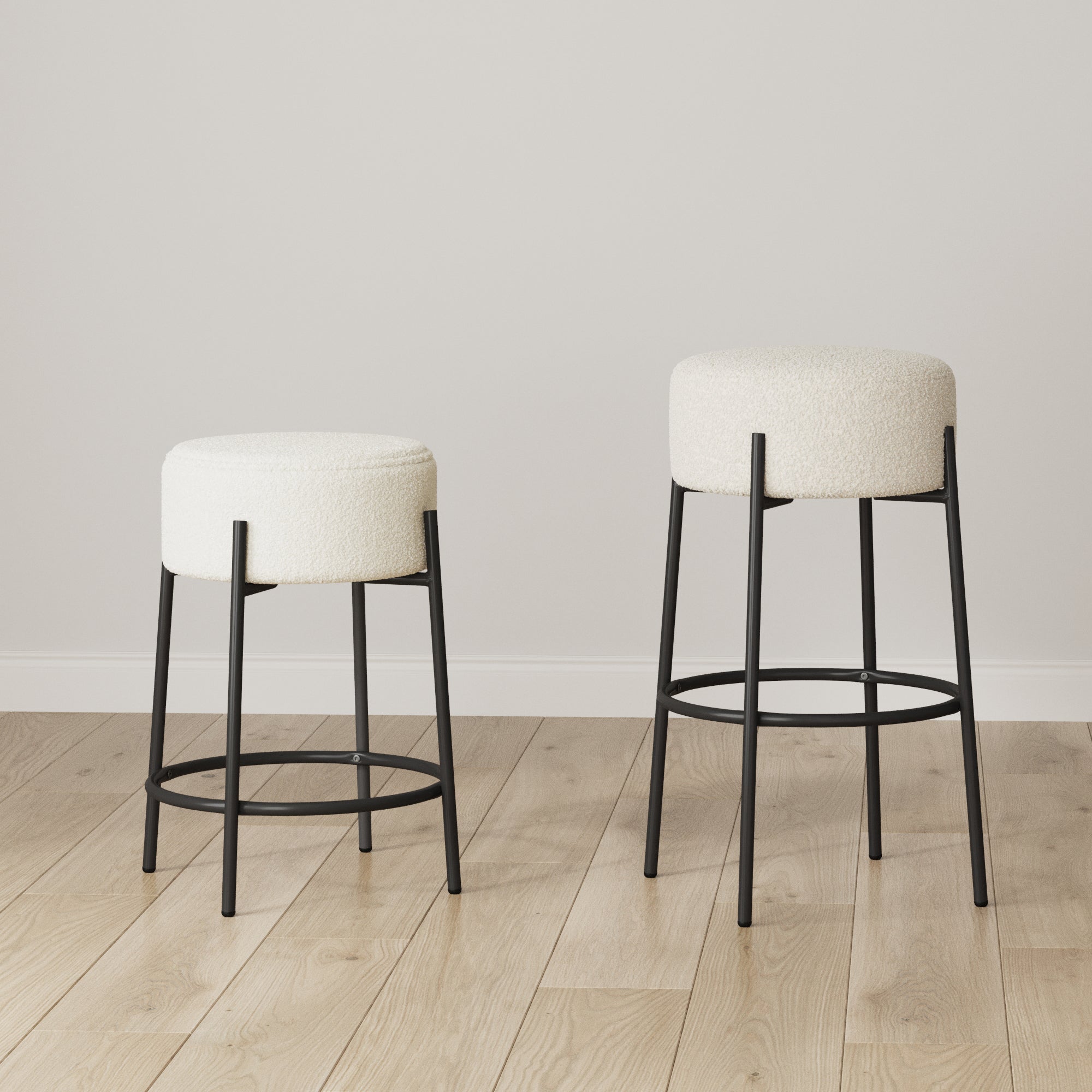 Two modern bar stools with black metal legs and round, white upholstered seats are placed on a light wood floor against a plain white wall. One stool is shorter than the other.