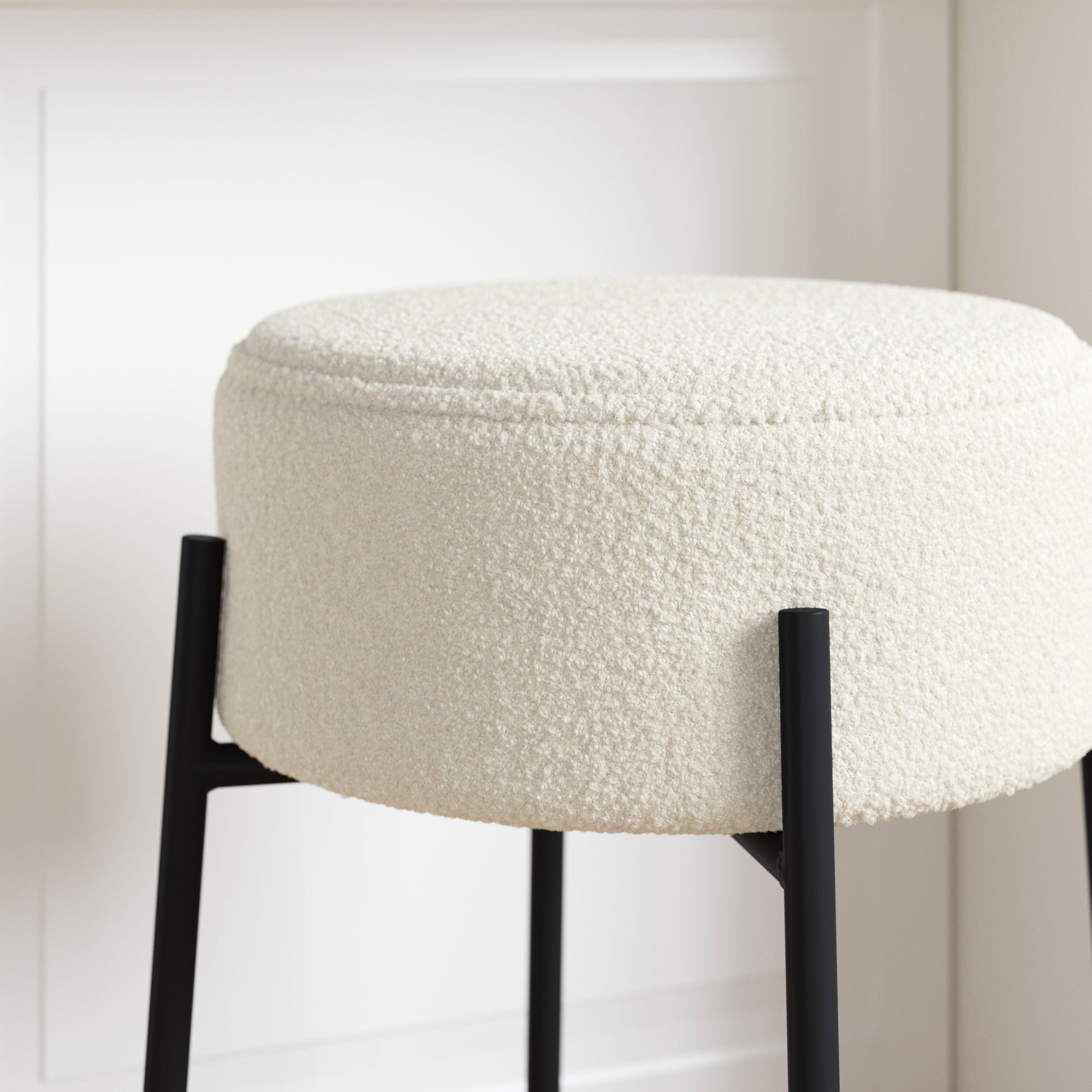 A round, white boucle fabric stool with black metal legs is positioned in front of a white paneled wall.