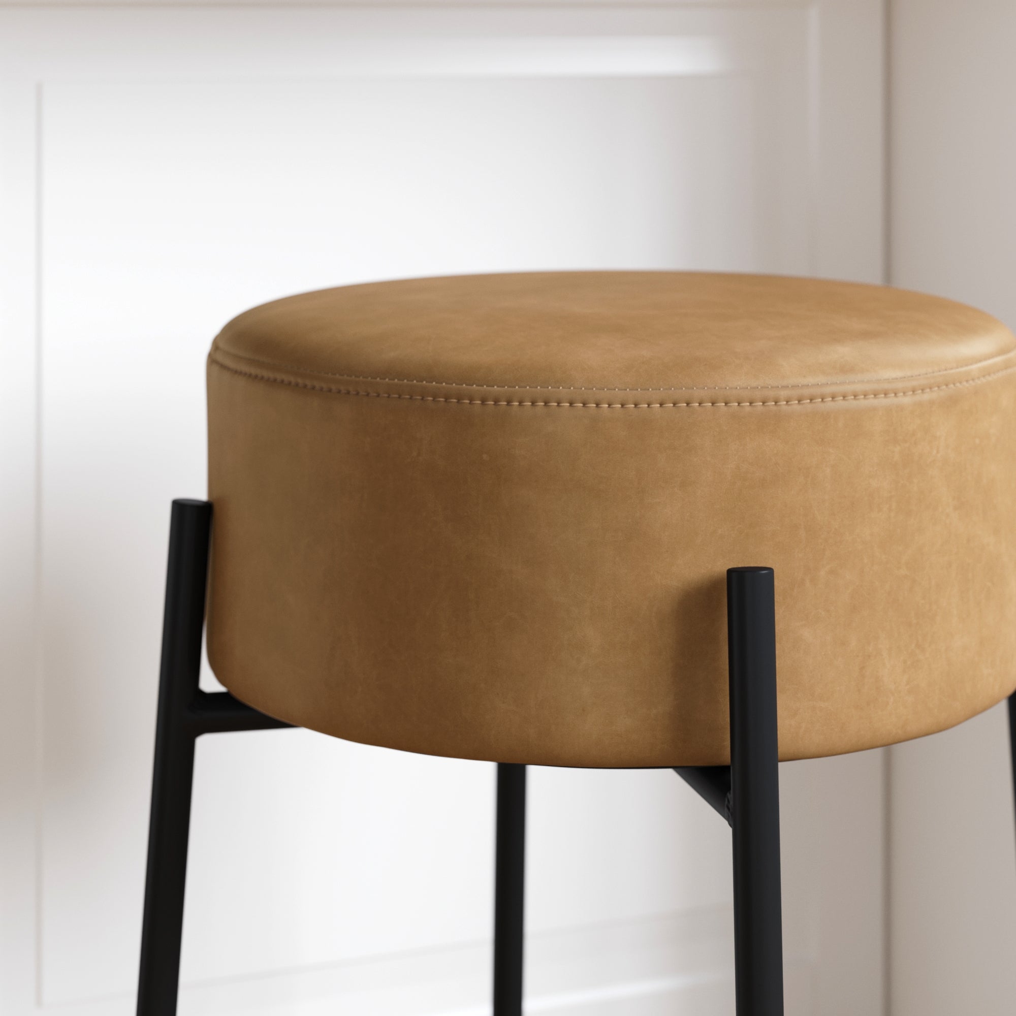 A round, tan leather stool with black metal legs is positioned against a white paneled wall. The stool has a smooth, upholstered seat and a modern, minimalist design.
