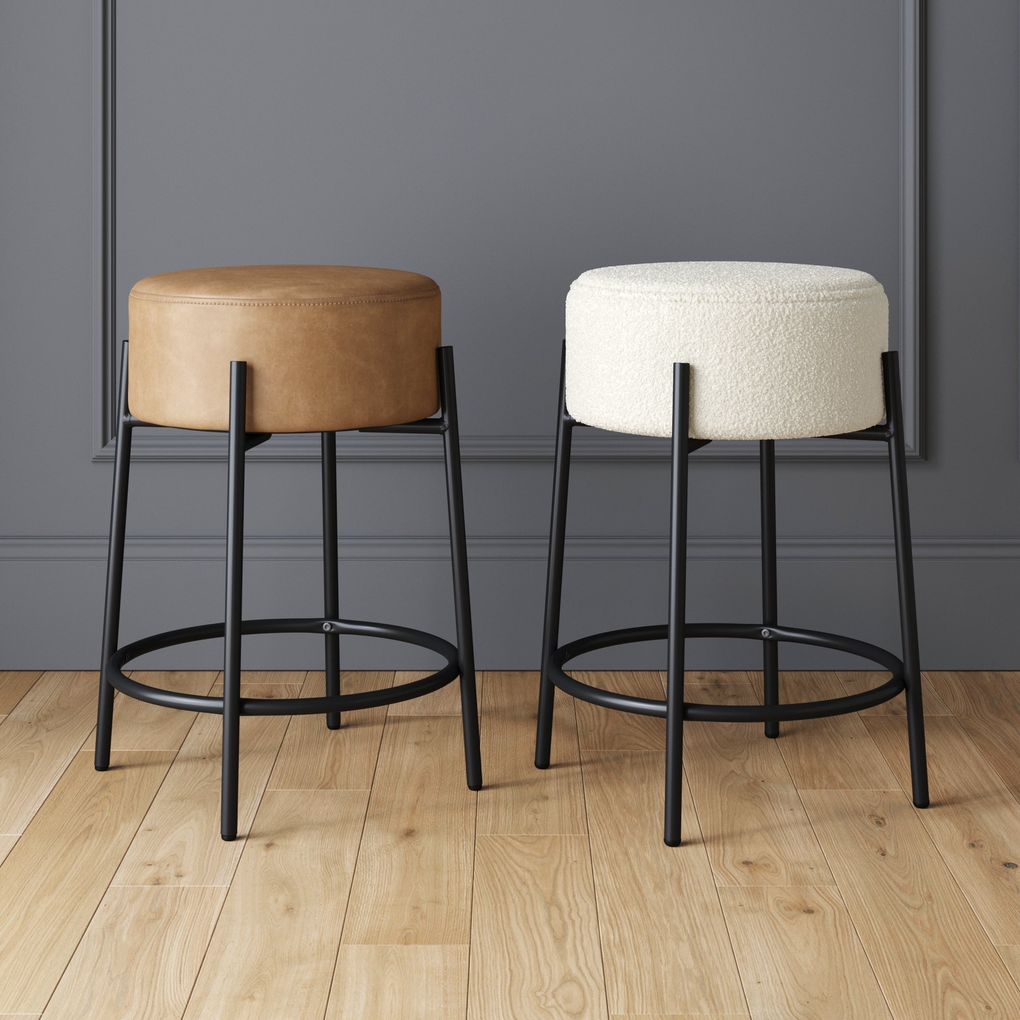 Two modern barstools with black metal legs; one has a brown leather seat, the other has a white textured fabric seat. They are placed on a light wooden floor against a gray wall with decorative molding.