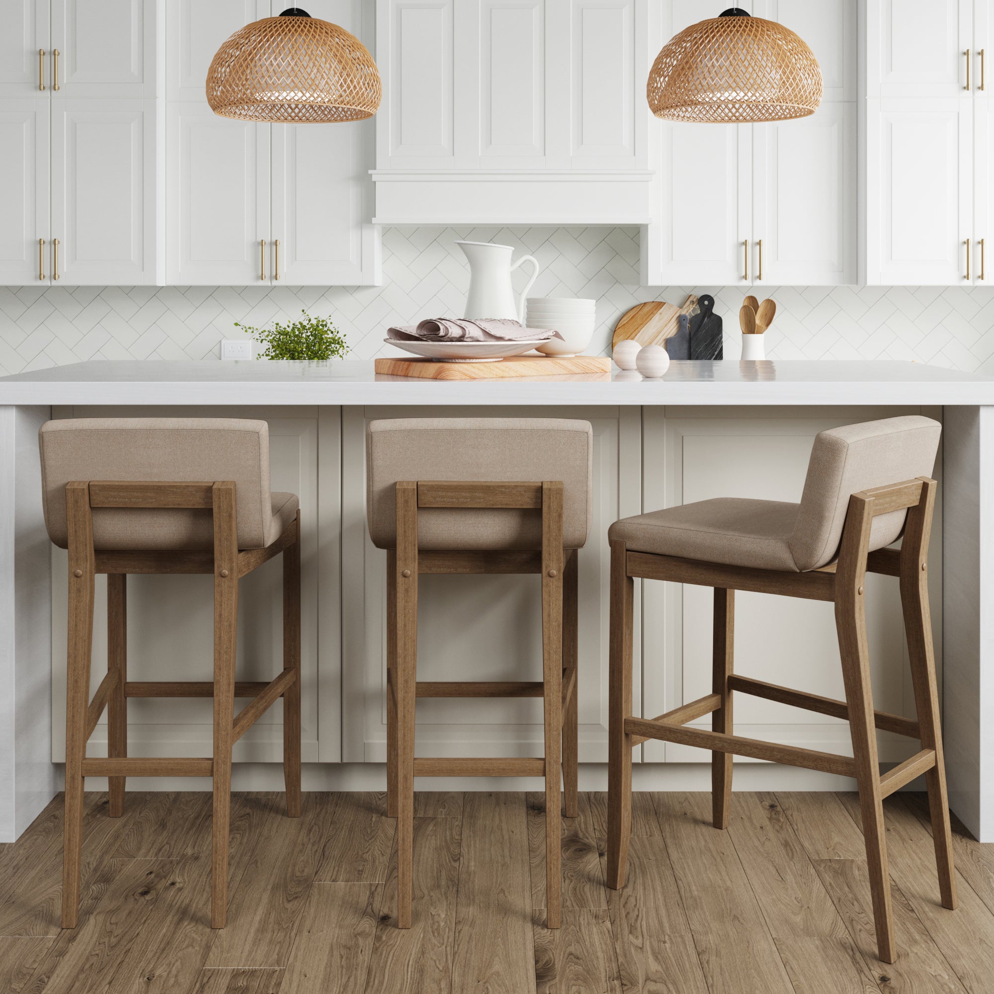 Modern kitchen with white cabinets, a white island, and three wooden bar stools with beige cushions. Woven pendant lights hang above, and kitchenware and plants decorate the countertop. Light wood flooring throughout.